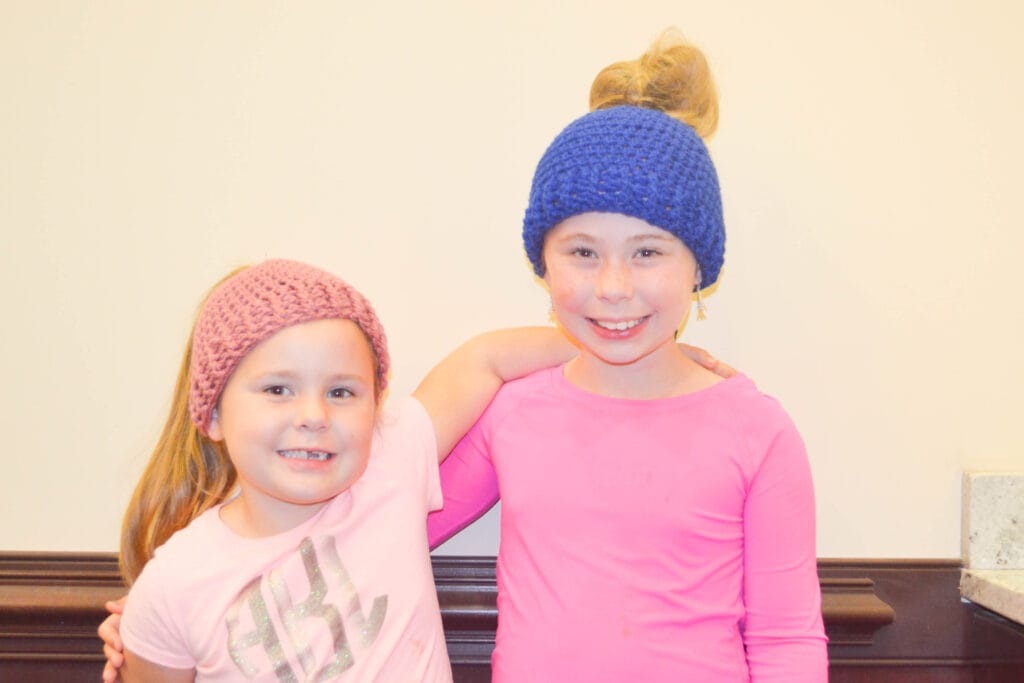 two girls are standing in front of a wall looking at the camera. One wears a pink crochet messy bun hat, while the taller girl wears a blue crochet messy bun hat.