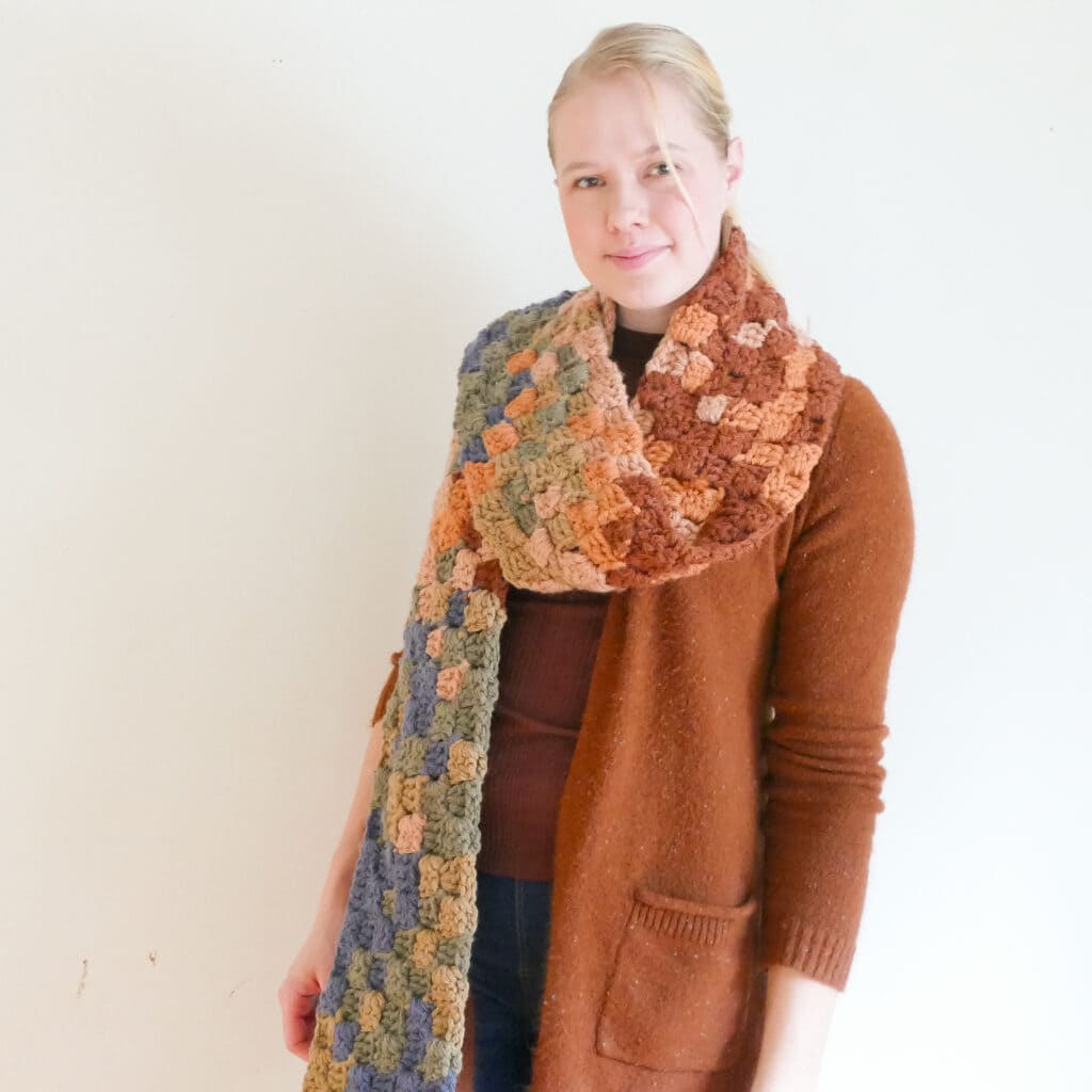 A blonde woman in a rust colored sweater against a light background showing a muliticolored corner to corner scarf done as a temperature project(temperature blanket alternative)